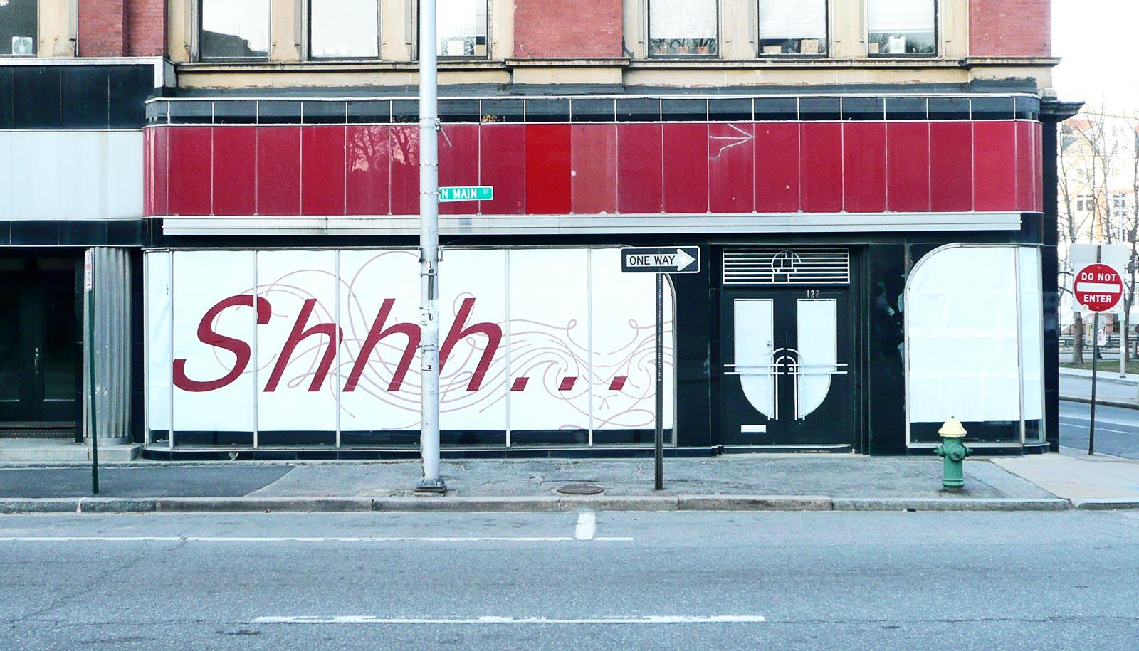 A building has "Shhh..." written in large letters as it stands on an empty sidewalk