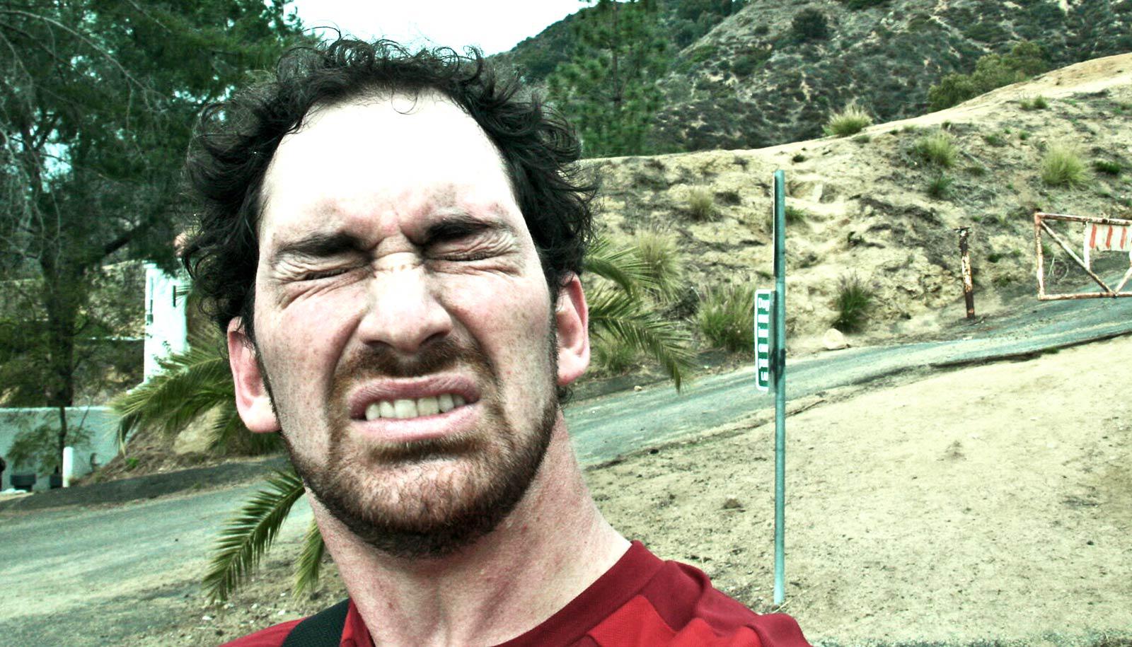 A man grimaces while sneezing outside on a walking trail