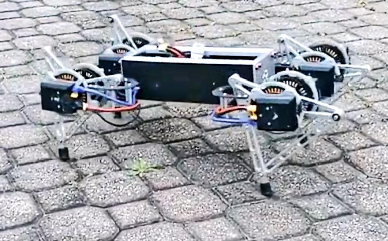 A four-legged robot stands on a outdoor path, with wires coming out of the main body and going into motors on the legs