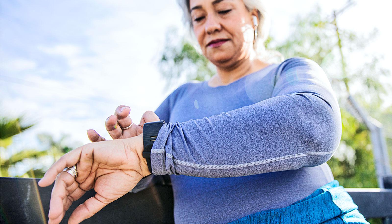 An older woman dressed in blue workout gear looks down to use her activity tracker, with a footpath railing behind her and the blue sky above