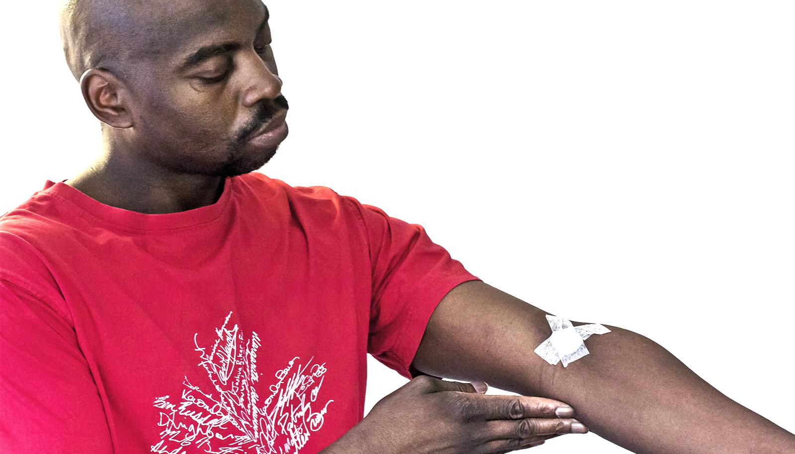 A man in a red shirt looks down at bandages covering where he's gotten a blood test