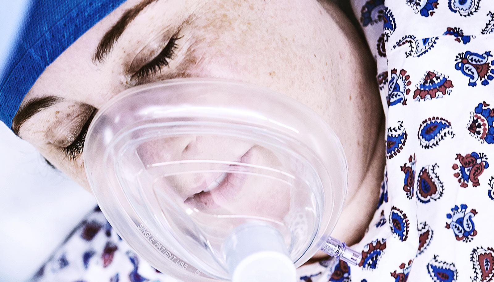 A woman in a hospital gown and with an anesthesia mask on is unconscious.