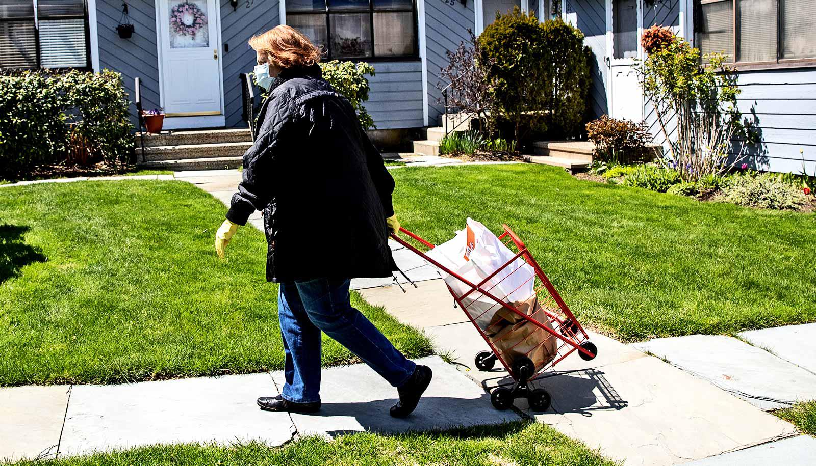 A woman pulls a cart filled with grocery bags down the sidewalk while wearing a mask and gloves