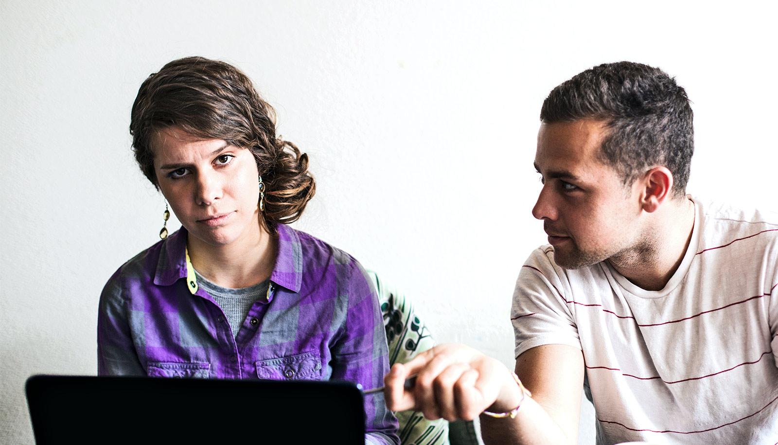 Two people sitting at a table, with one talking to the other and one looking at the camera, exasperated