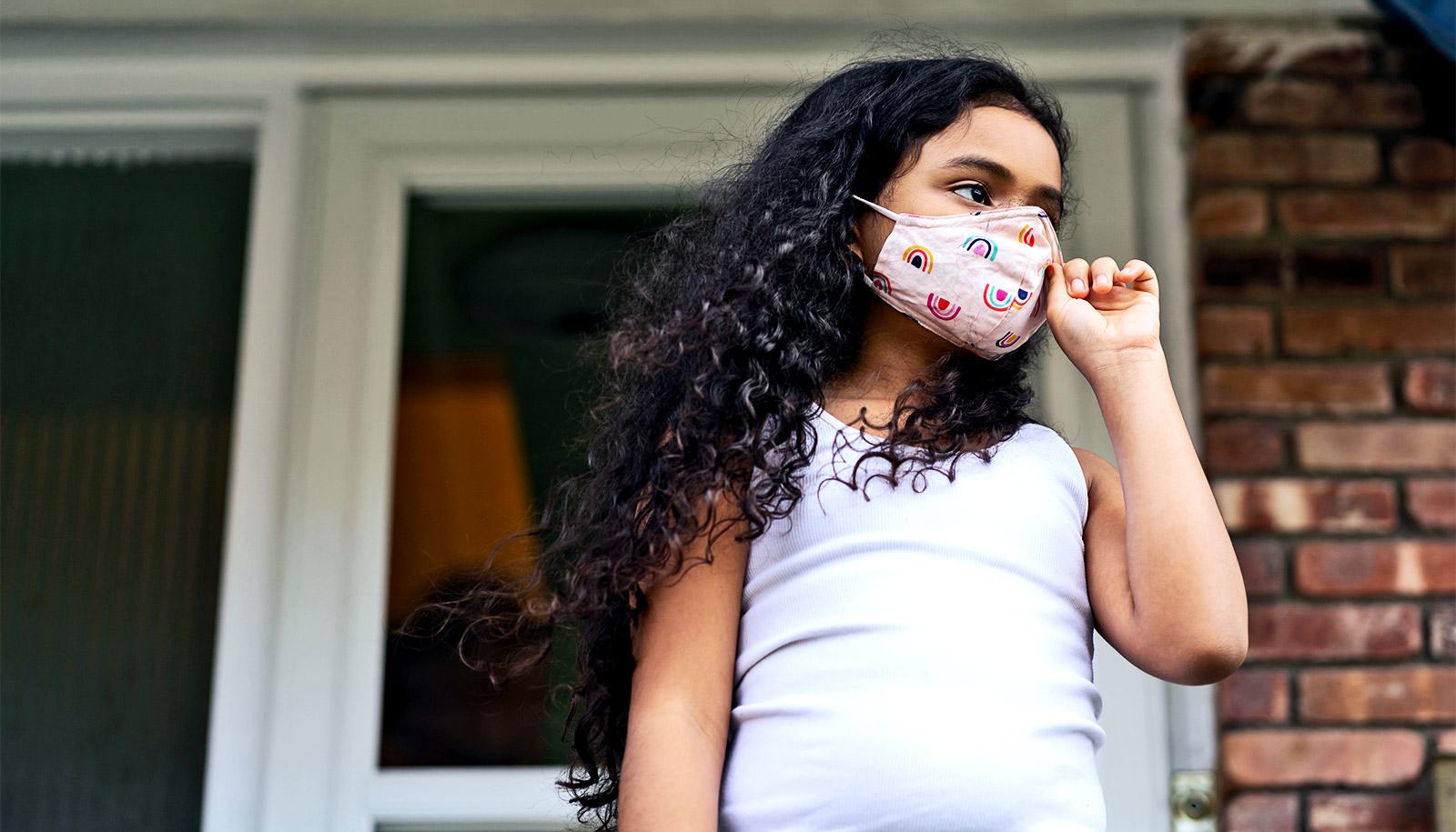 A young girl wearing a face mask stands outside her house looking up the street