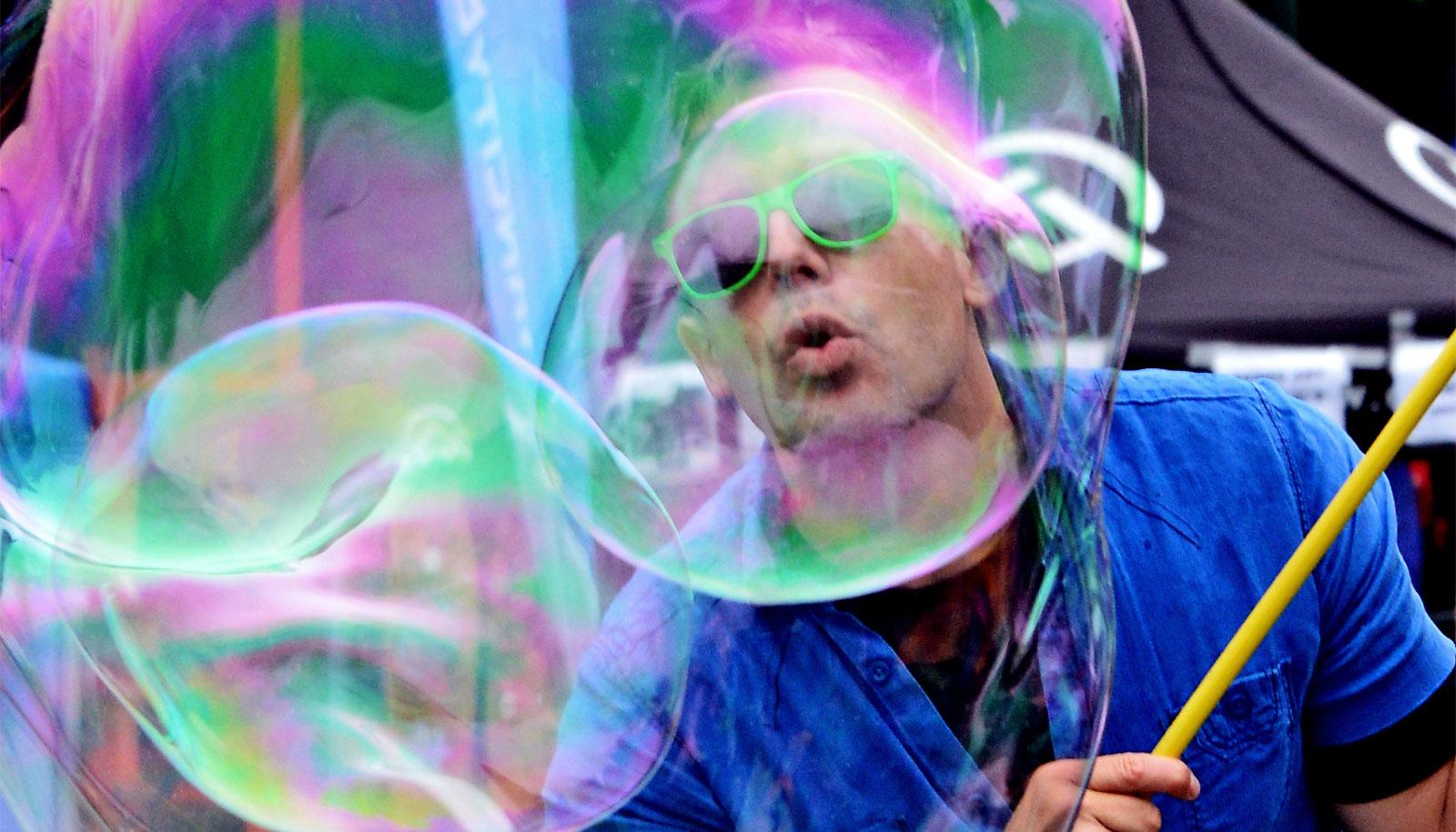 A man wearing a blue shirt and green sunglasses creating a giant soap bubble blows into the bubble