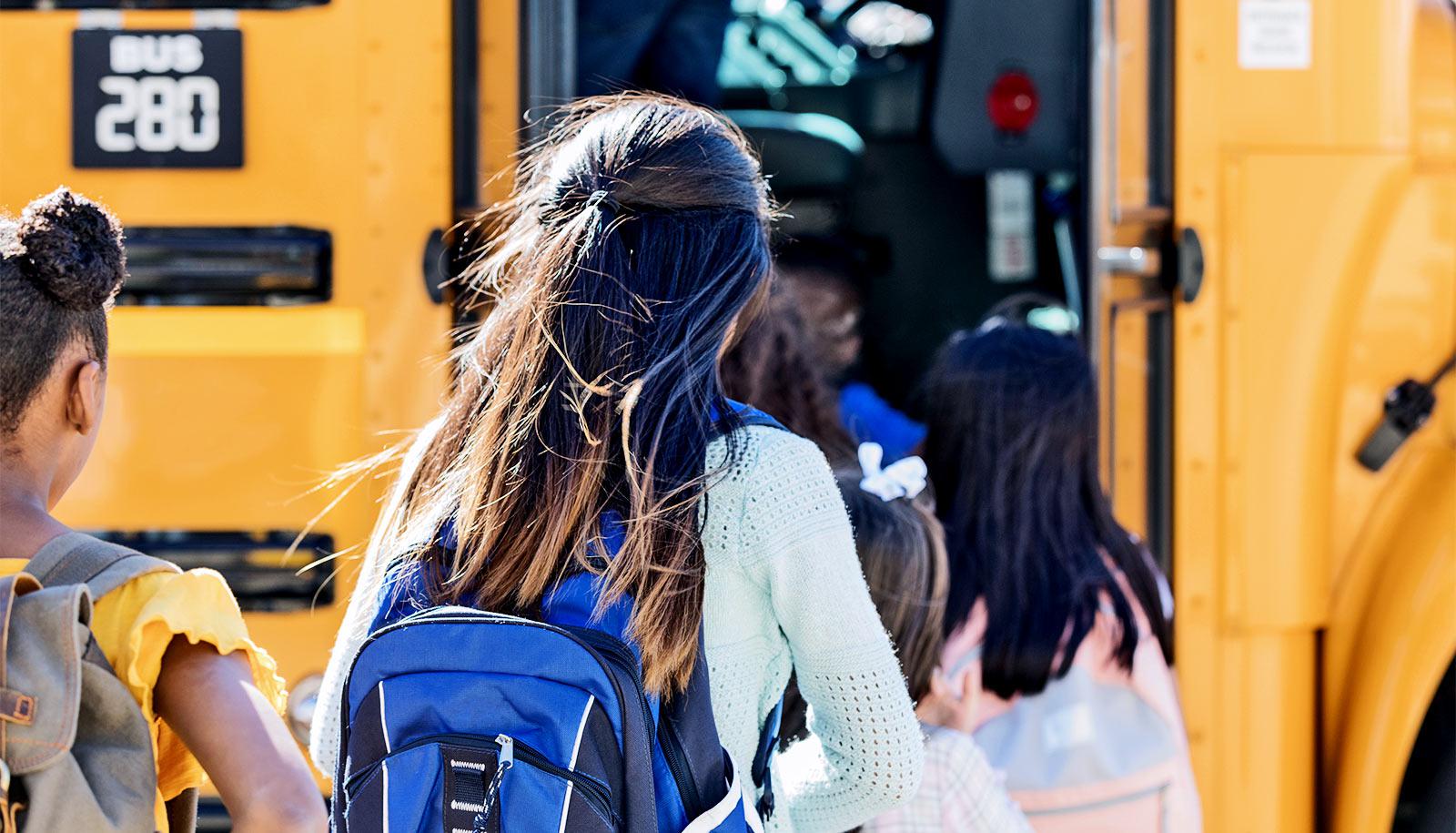 A young girl in a blue sweater with a blue backpack looks down as she and a line of middle school students file onto a school bus