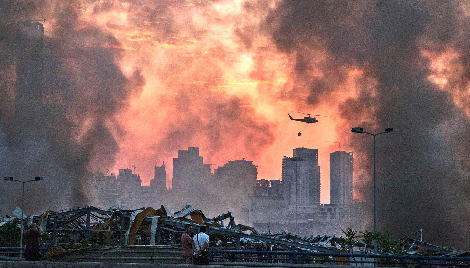 Helicopters fly over the site of the explosion as people look over wreckage and smoke billows into an orange sky