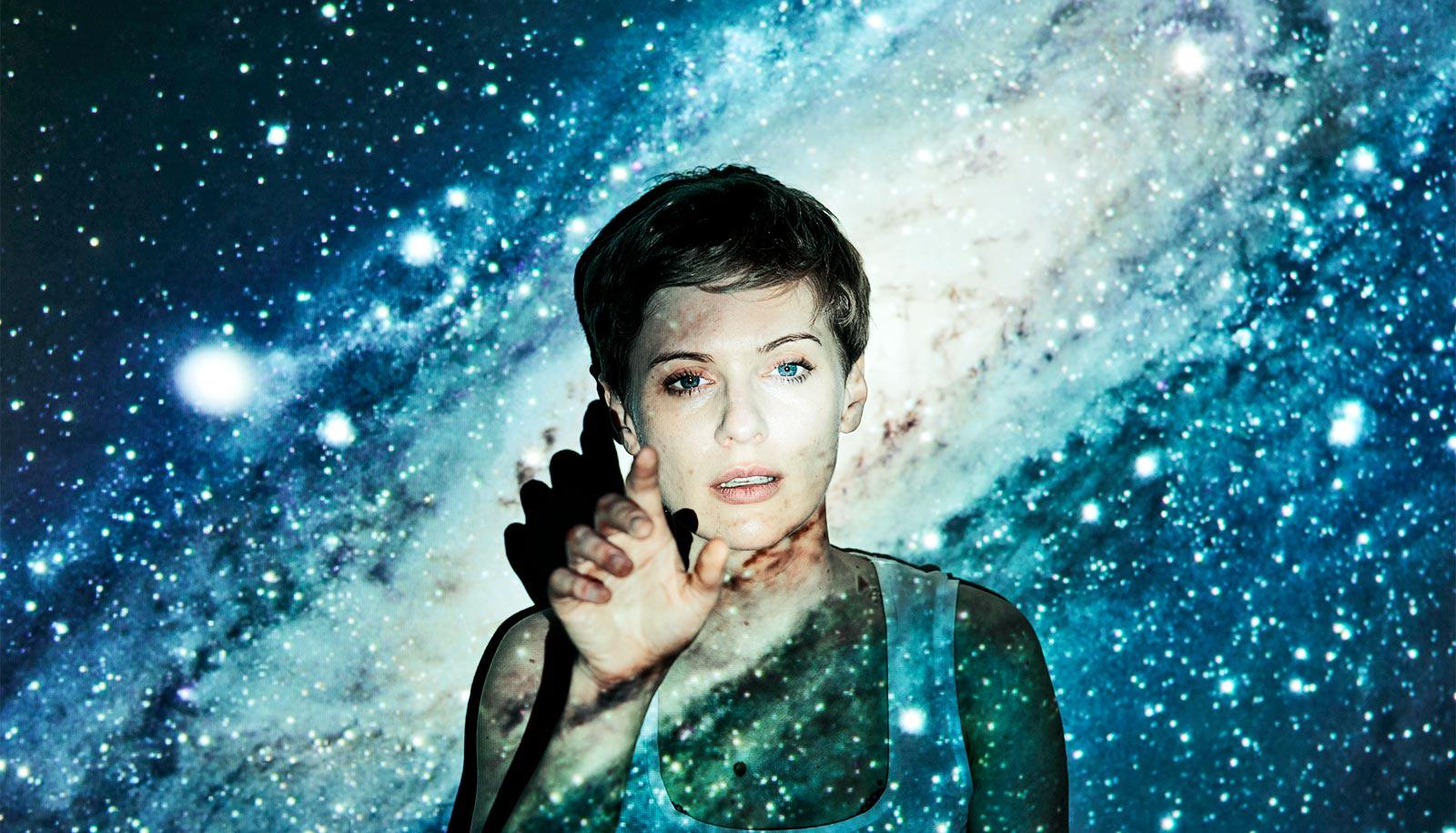 A young woman looks curious as she reaches her hand out while an image of space is projected on her face