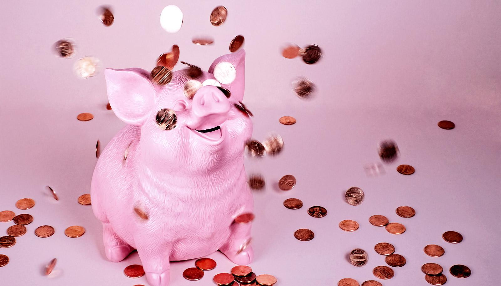 A piggy bank sits on a pink background with coins raining down on it. It's smiling face is looking up as the coins fall down