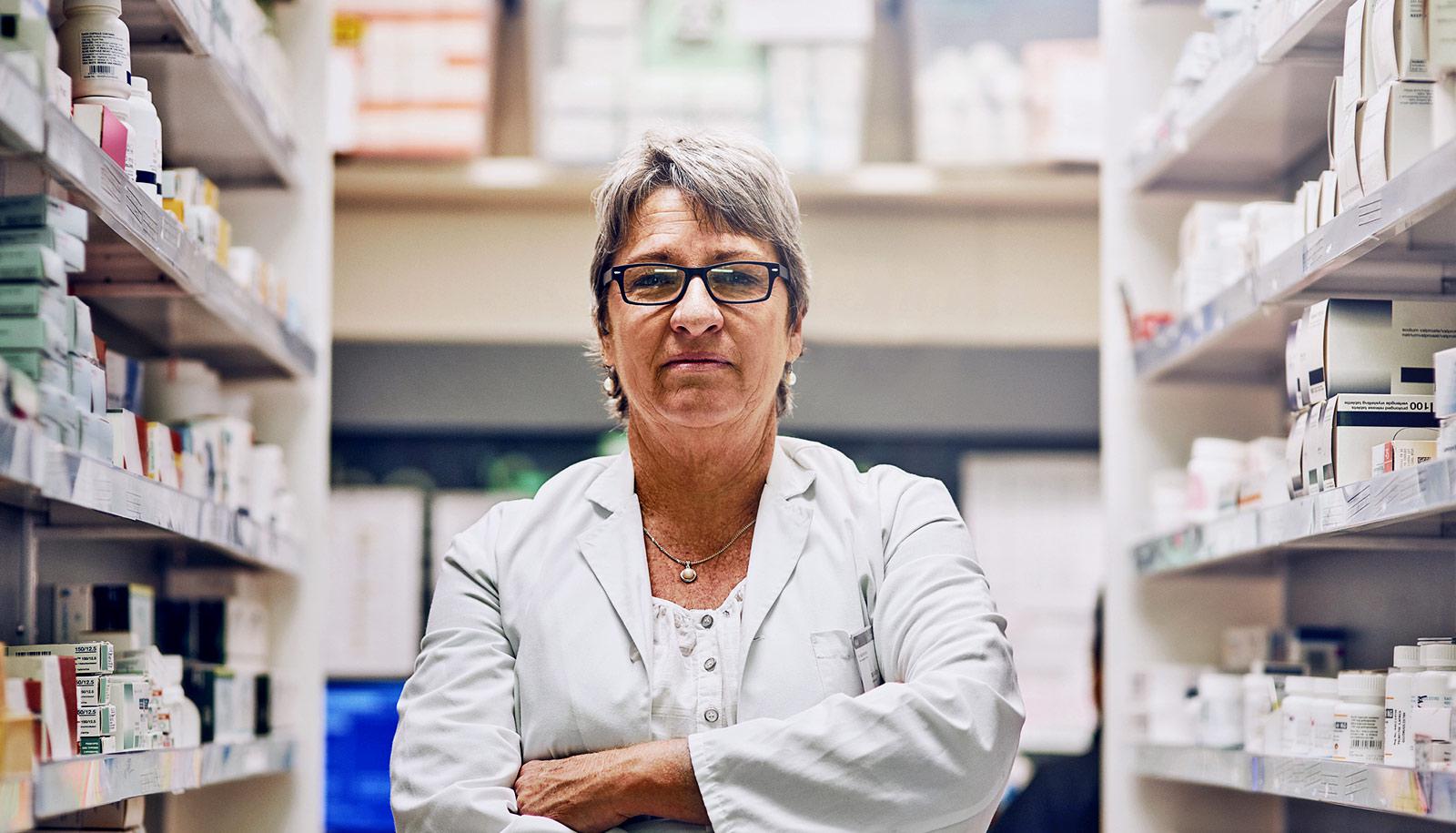 serious pharmacist in white coat crosses arms between two shelves full of medications