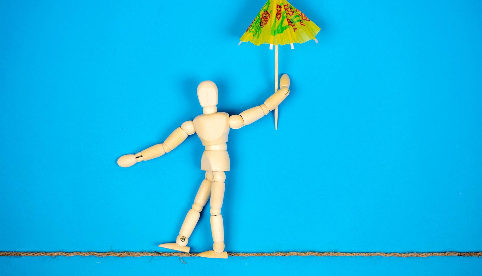 A wooden anatomy figure looks like its walking a tightrope while holding a cocktail umbrella