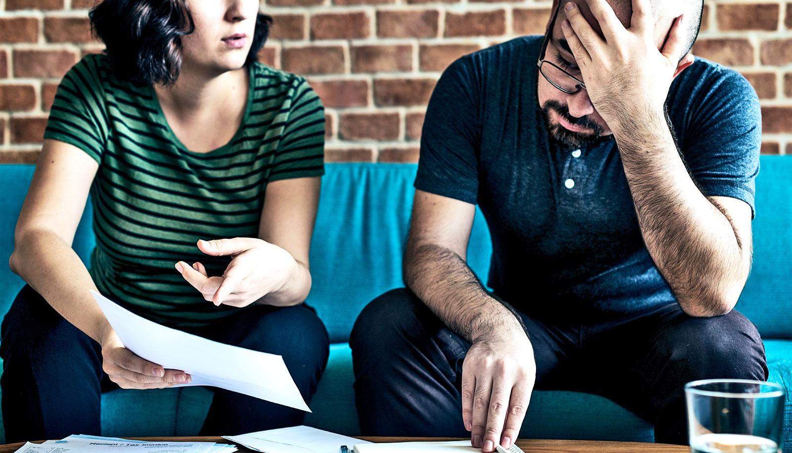 A man and woman look at their bills, as she points to a piece of paper and he puts his head on his hand